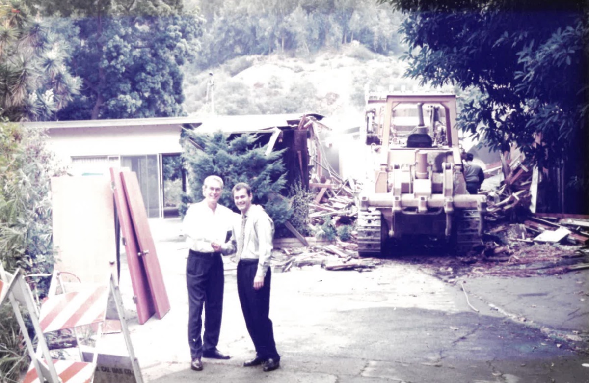 Two men in shirtsleeves shaking hands on a driveway in front of a single-storey house being pulled down behind them, a bulldozer at work in the rubble.