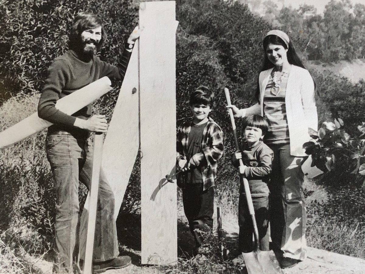 A black-and-white family photograph: a bearded man holding a surveyor’s stake beside a woman and two children carrying tools, on an undeveloped hillside lot.