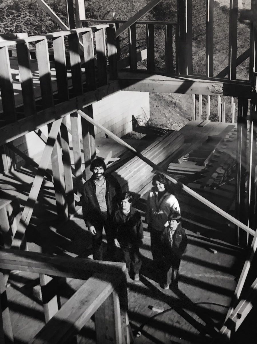 A black-and-white photograph looking down through the open timber frame of a house under construction, a family of four standing together on the deck below among stacked lumber.